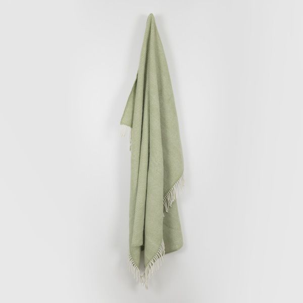 The Fern Green Fishbone Throw hanging up on a hook on a plain grey background.