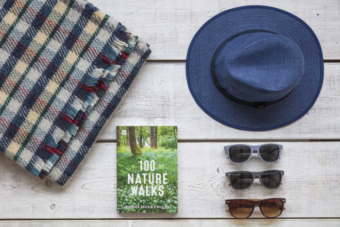 Top down view of the Recycled Rug folded up laying on a wooden background alongside a blue ambassador hat, three pairs of sunglasses and a book called 100 nature walks.