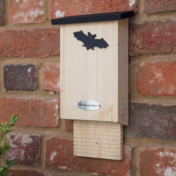 Wooden bat box on a red brick wall. The bat box features a National Trust metal logo on the front and a bat silhouette cut out. The FSC logo is visible on the side.