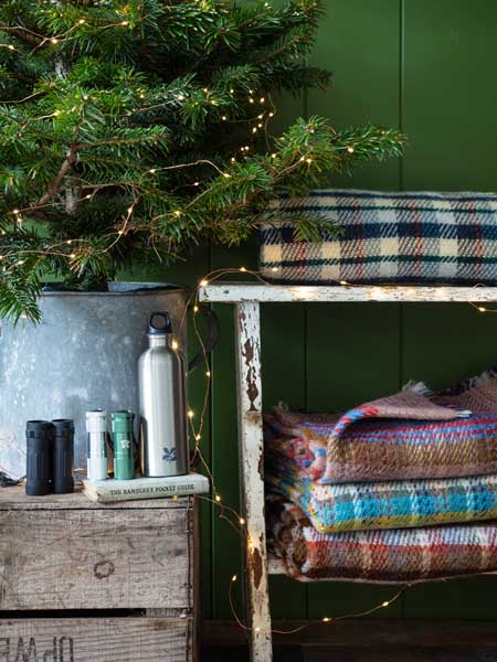 National Trust outdoor accessories range, great gifts for outdoor explorers. Image shows Recycled Rugs in varying colours stacked under a real Christmas tree, with walking and hiking accessories on bench nearby. Browse the range for gifts that help look after National Trust places, 365 days a year.