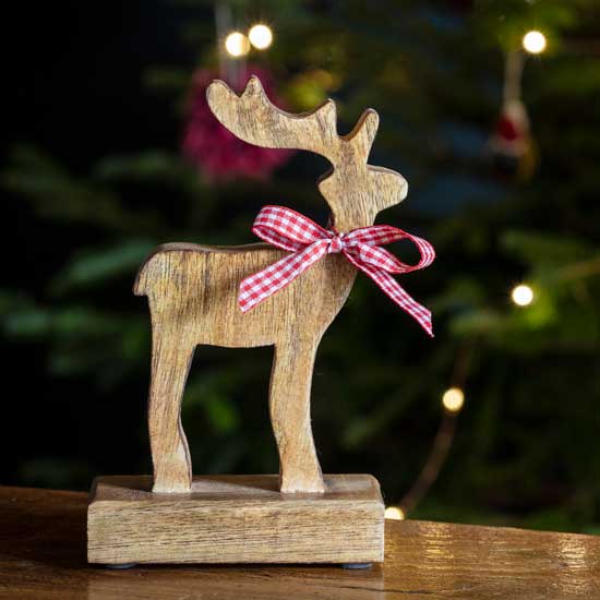National Trust Wooden Reindeer with Bow Decoration, image shows wooden sculpture on sideboard with Christmas tree in background. Part of the National Trust Christmas decorations range, browse the Christmas collection for sustainably made gifts that help look after National Trust places.