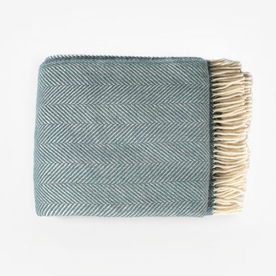 Herringbone Wool Throw – Petrol Blue. Image shows wool throw folded with cream tassels and blue herringbone weave on a plain grey background.