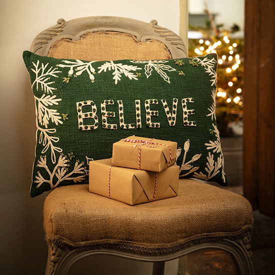 Believe Cushion - Embroidered, image shows forest green oblong cushion on a vintage chair accompanied by neatly wrapped gifts.