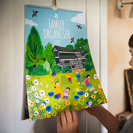 National Trust Family Organiser 2025 - image shows calendar hung against a larder cupboard door, with person lifting the cover to add details to an inner page.