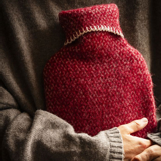 Hot Water Bottle and Cover, Red - image shows hot water bottle with red woollen cover and cream blanket stitching held in arms of person.