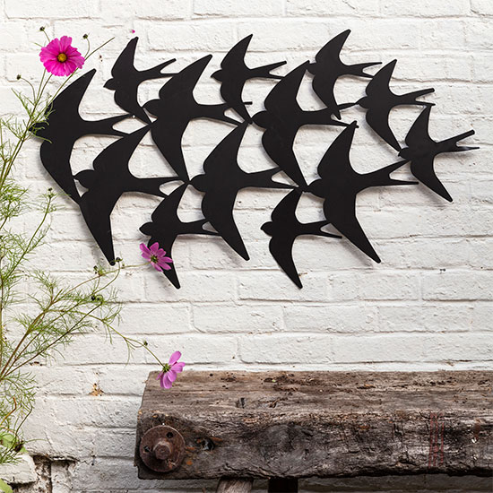 Swallows Garden Wall Ornament. Image shows large metal garden ornament in the shape of migrating swallows hung on a white garden wall, with a rustic bench beneath and green foliage. 