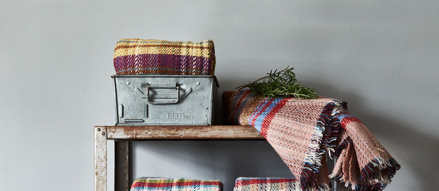 Woven with history - Made in the UK by home textile experts, browse our range of quality throws and blankets for homeware that helps look after the special places in our care. Image shows several of the National Trust Recycled Rug in varying colourways and patterns folded in storage baskets, arranged on metal industrial style shelving in a neutral coloured hallway.