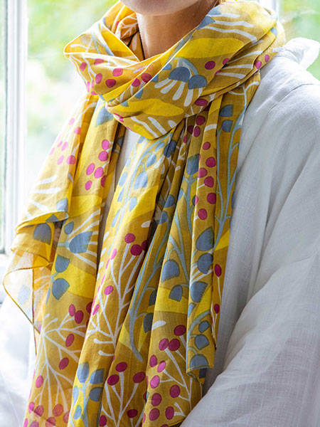 Fashion accessories - browse the range for scarves, bags, jewellery and more. Designed with nature in mind, browse the National Trust fashion range and help look after the places in our care with every purchase. Image shows Lily of the Valley Scarf in bright, uplifting yellow worn on model. Made from 100% organic cotton, this scarf features a grey, white and red print on a bold yellow fabric.