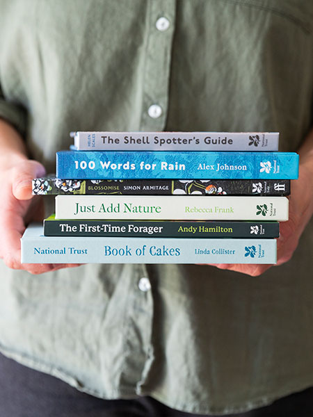 Books. Image shows selection of National Trust books, including Blossomise and Book of Cakes, held in hands by model wearing jeans and grey green shirt.