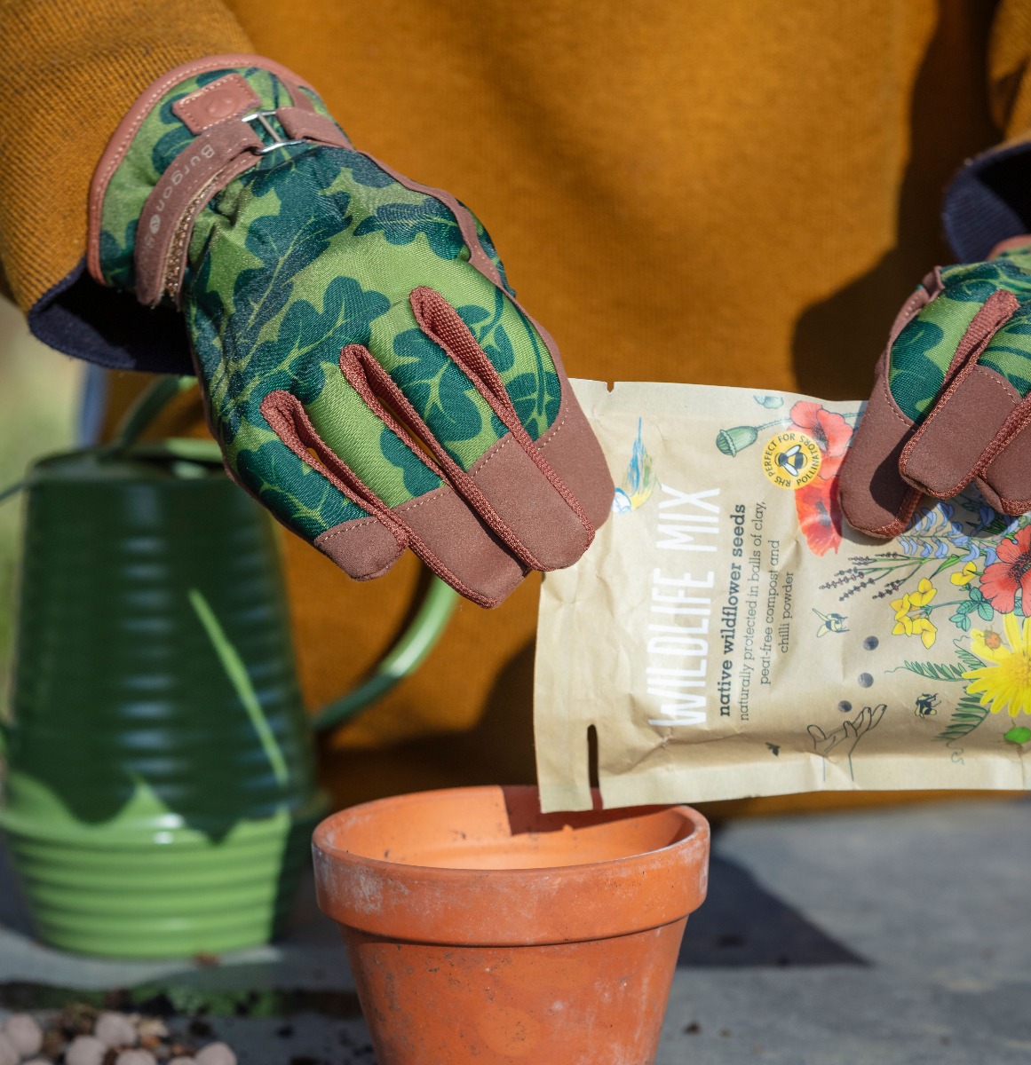 Moss Oak Leaf Gardening Gloves (£18.00), from the National Trust's gardening range. Image shows gardening gloves worn by gardener, planting seeds on a garden workbench. Every purchase from the National Trust shop helps care for nature, beauty and history for everyone, for ever.
