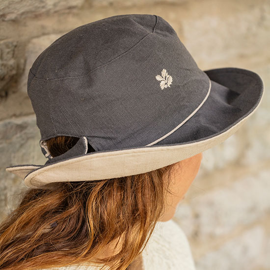 Reversible Linen Hat(£16). Image shows hat in dark grey worn by model sat in garden. Hat features a lighter beige underneath, with embroidered National Trust oakleaf on the main body of hat.