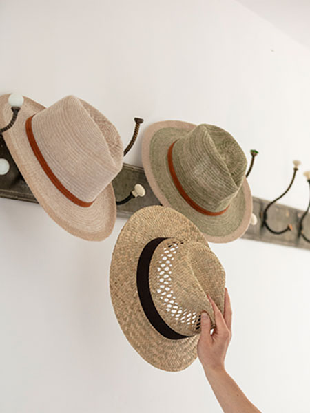 Fashion accessories. Image shows hats hung on vintage hooks, including our National Trust Straw Hat with Band and Powder Ivory Cotton Hat, with a hand reaching towards hats.