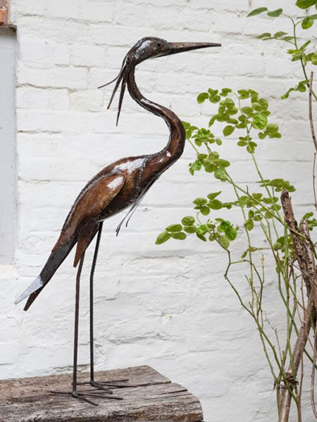 Garden decor. Image shows Metal Heron Sculpture on a garden table, surrounded by green foliage.