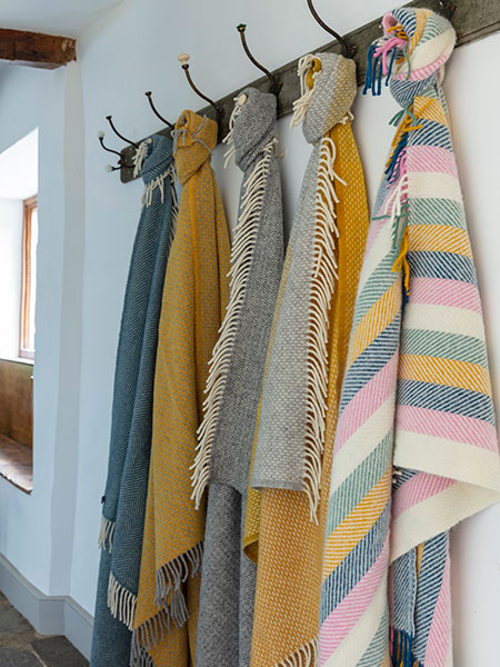 National Trust throws and blankets range - image shows several wool throws, including our new Coastal Sunset and Fishbone Stripe colourways, hung in a hallway on vintage style hooks. Browse homeware inspired by nature, beauty and history - every purchase purchase helps look after nature, beauty and history.
