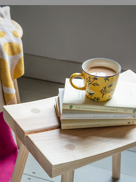 Mugs, cups & ceramics - browse the new spring homeware collection for mugs, blankets, cushions and more inspired by nature. Image shows Cowparsley Mustard Mug sat atop an Ebworth Curved Stool, with Jacquard Leaves Throw in the background.