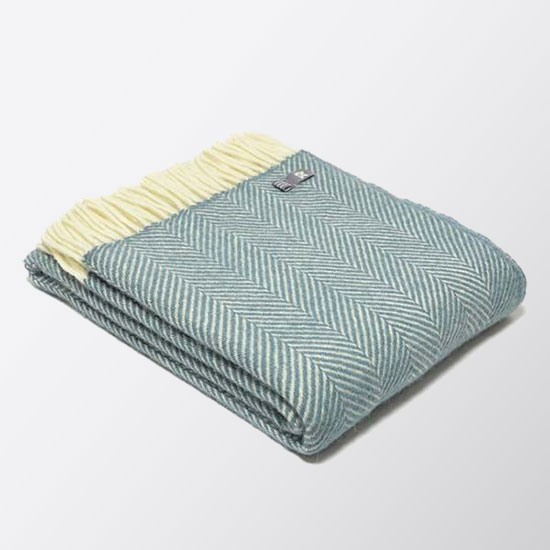 National Trust Wool Blanket, Petrol Blue (£48.00). Image shows woollen throw, with a fishbone weave pattern, in a mid blue shade. The blanket is folded, displayed on a plain grey background. 