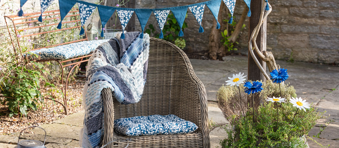 Image shows a variety of products from the garden decor and outdoor living range in a garden setting, with the Bluebell Bunting and Bluebell Seat Pad, as well as the Daisy and Cornflower Plant Stakes, decorating the scene. The copy reads: Sunshine ready - Give the garden a fresh new look with unique sculptures and decorations, from soft furnishings and solar powered lighting to flora and fauna inspired plant stakes.<