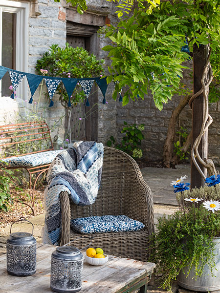 Outdoor living. Image shows a variety of garden accessories in a garden setting - from National Trust seat pads and bunting in our new 'Bluebell' design, solar outdoor lanterns and more, arranged around a wooden pergola and wicker garden chair.