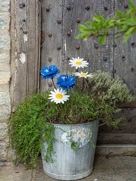 Plant Stakes - browse the garden range for plant care, wall art, sculptures and more. Designed with nature in mind, browse the National Trust garden collection and help look after the places in our care with every purchase. Image shows Cornflower Plant Stake and Daisy Plant Stake in a metal planter, surrounded by green foliage. Made from recycled metal, these plant stakes are both decorative and functional - give your plants a helping hand, while brightening up your outdoor space.