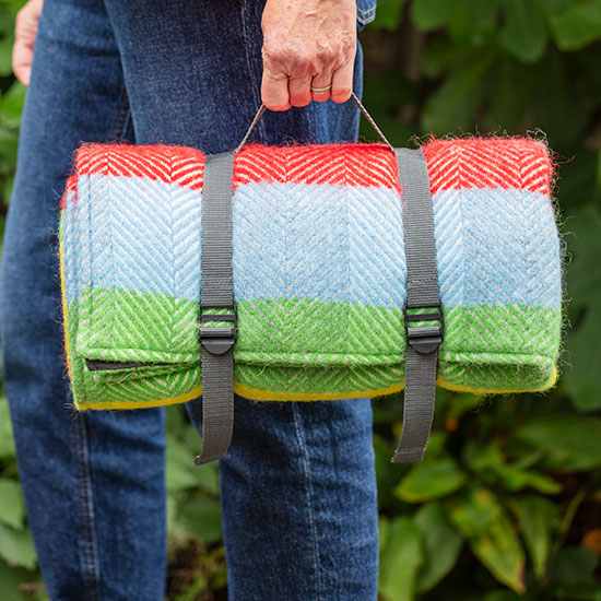 Waterproof Backed Rainbow Picnic Rug (£45.00). Image shows picnic rug, with rainbow coloured stripes, rolled and carried by model in garden.
