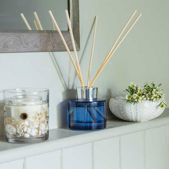 Reed Diffuser - Bergamot and Seamoss (£22). Image shows reed diffuser, with blue glass container and FSC reeds, on shelf in bathroom.
