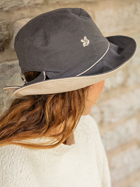 Outdoor accessories. Image shows Reversible Hat in Grey Beige worn by model, sat in a garden setting.
