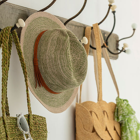 Powder Green Sun Hat(£30). Image shows sun hat in light green hung on vintage hooks in country house. Also hung on hooks nearby are the Ellya Sea Grass Tote Bag and Green Seagrass Bag with Handles from the wider National Trust fashion accessories range. 