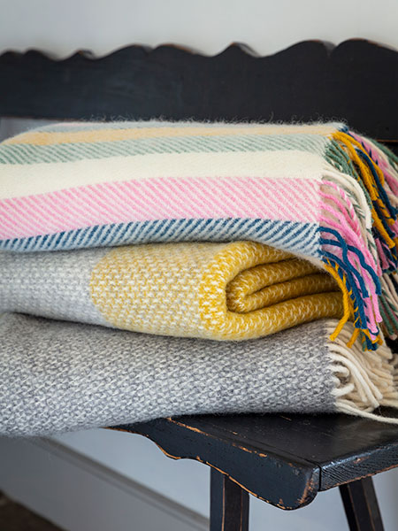 National Trust throws and blankets range - image shows several wool throws, including our new Coastal Sunset and Fishbone Stripe colourways, folded and stacked on a vintage bench. Browse homeware inspired by nature, beauty and history - every purchase purchase helps look after nature, beauty and history.