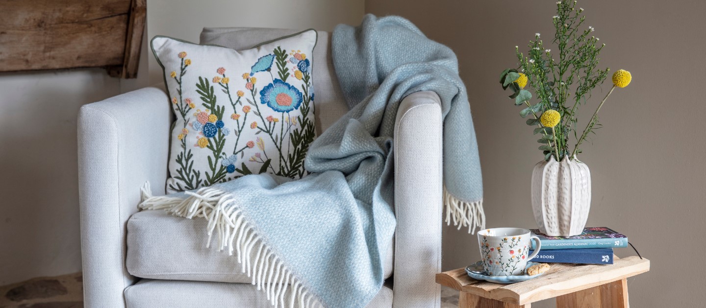 Spring summer collection - Discover our spring summer collection, bursting with unusual gift ideas, unique homeware and fashion favourites inspired by the places in our care. The image shows a cosy and inviting interior setup. It features a light beige armchair with wooden legs, adorned with a National Trust embroidered cushion featuring colourful floral patterns. Draped over the armchair is a soft National Trust duck egg throw with white fringed edges. Next to the armchair is a small wooden stool with a natural, rustic finish. The stool holds a taupe pod vase filled with fresh greenery and yellow billy button flowers, alongside a green wild daisy mug on a saucer, a biscuit, and a stack of books. The scene is set on a stone floor, enhancing the rustic charm of the space.