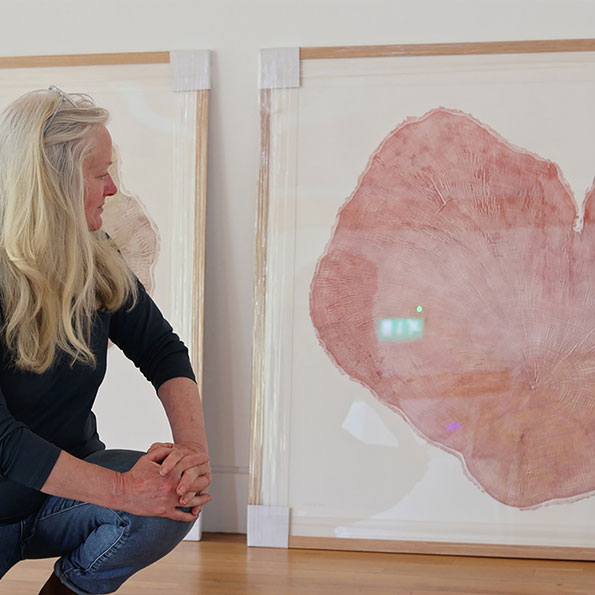 Remembering Sycamore Gap - prints by Shona Branigan from her original artwork Heartwood, a selection of reliefs taken from the Sycamore Gap tree trunk, comissioned by the National Trust. Image shows Shona bedside a lifesize print of Heartwood in red. 