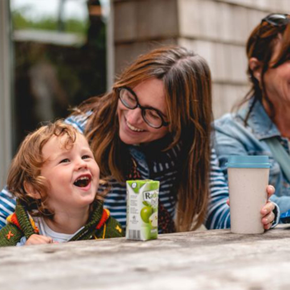 National Trust cafe free hot drink with every Circular & Co reusable cup promotion.  Image shows adult and child sat outdoors, at a garden table enjoying food and drink. Adult holding Circular & Co reusable cup, in light blue and cream.